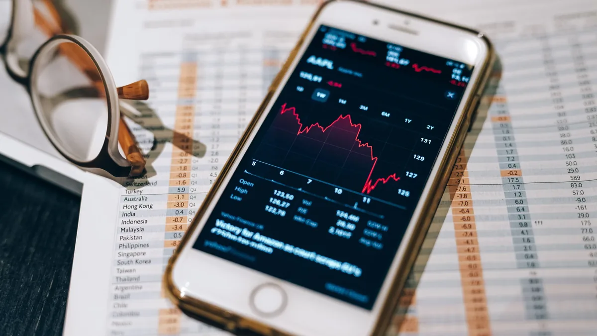 A smartphone displaying a stock market graph on its screen, placed on top of a document with a list of countries and numbers, accompanied by