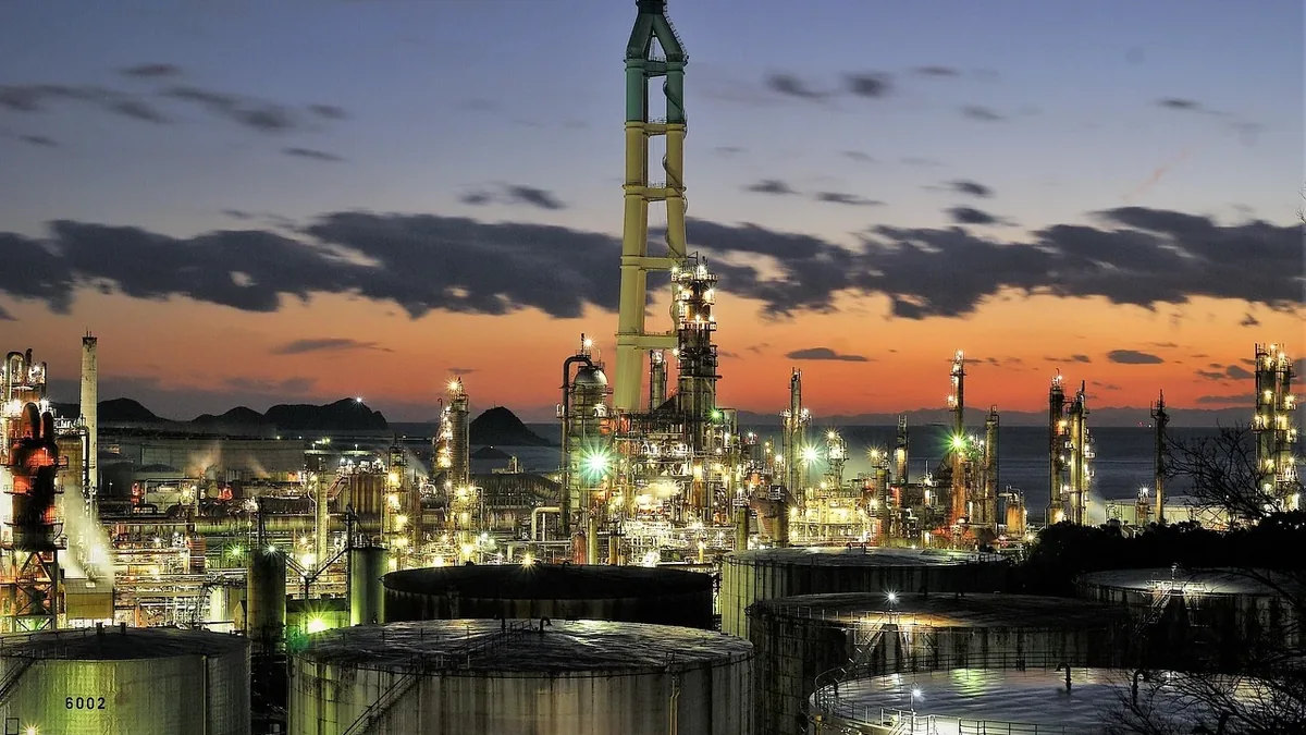 A large industrial complex with numerous tall structures, likely oil refineries or chemical plants, set against a sunset sky with clouds.